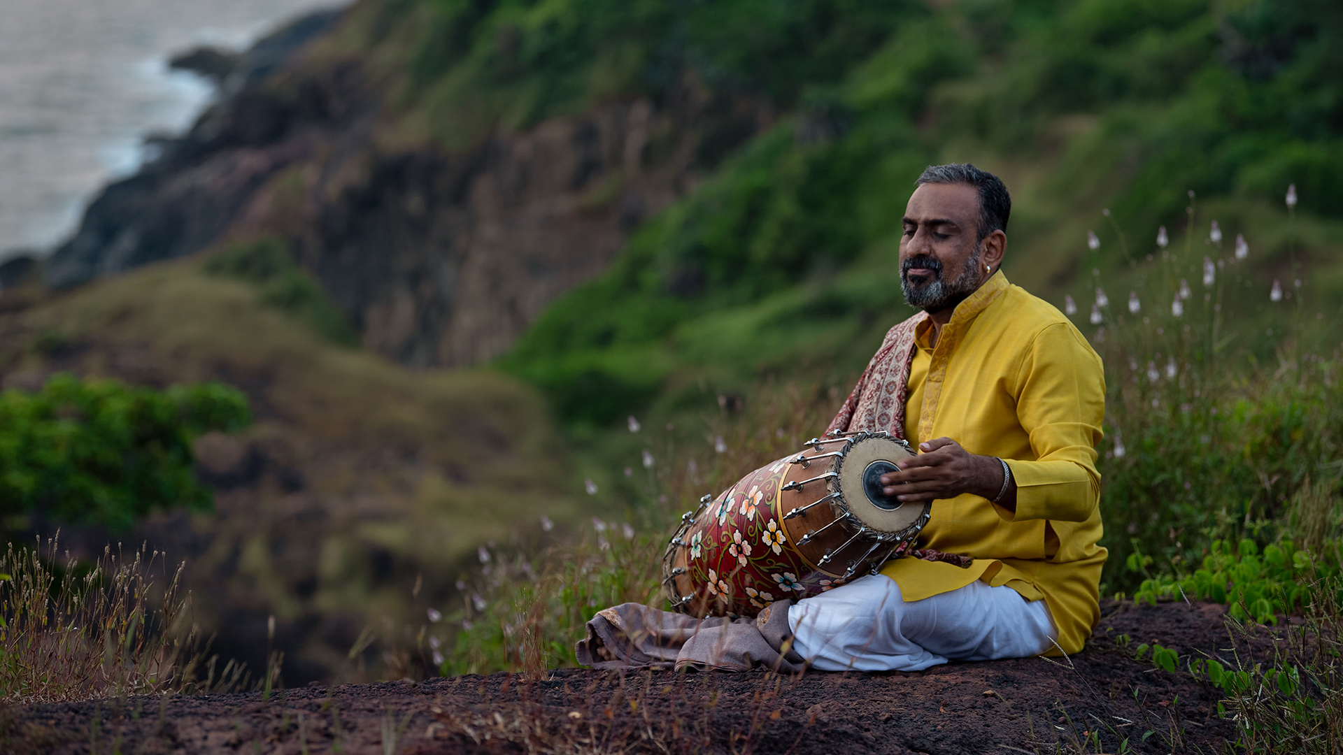 Jayachandra Rao playing the mridangam on a grassy clifftop above the sea at dusk.